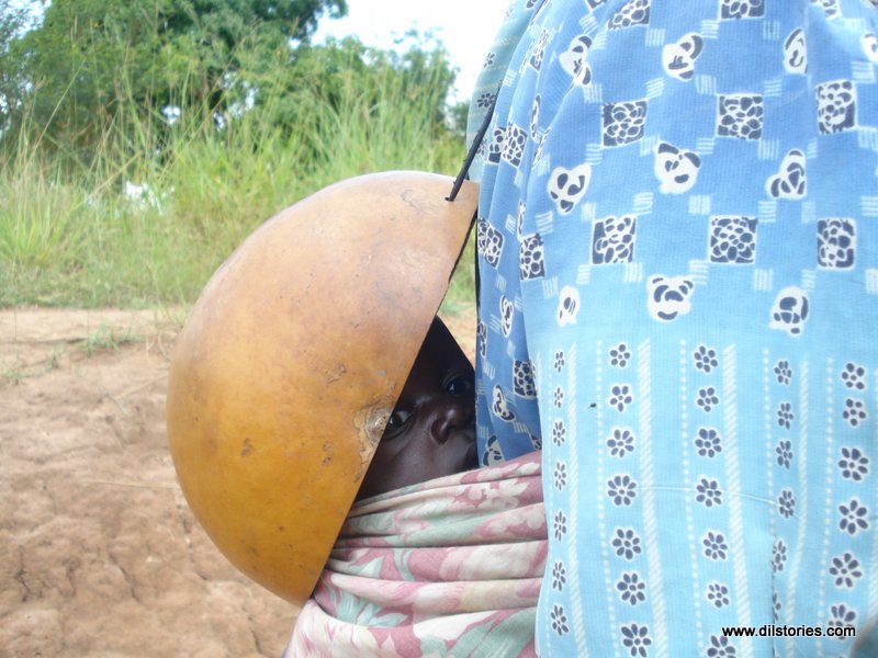 A child on the mother's back, a calabash protecting it from the hot sun. Mother in a blue dress, uses pink shawl to tie baby to her back.
