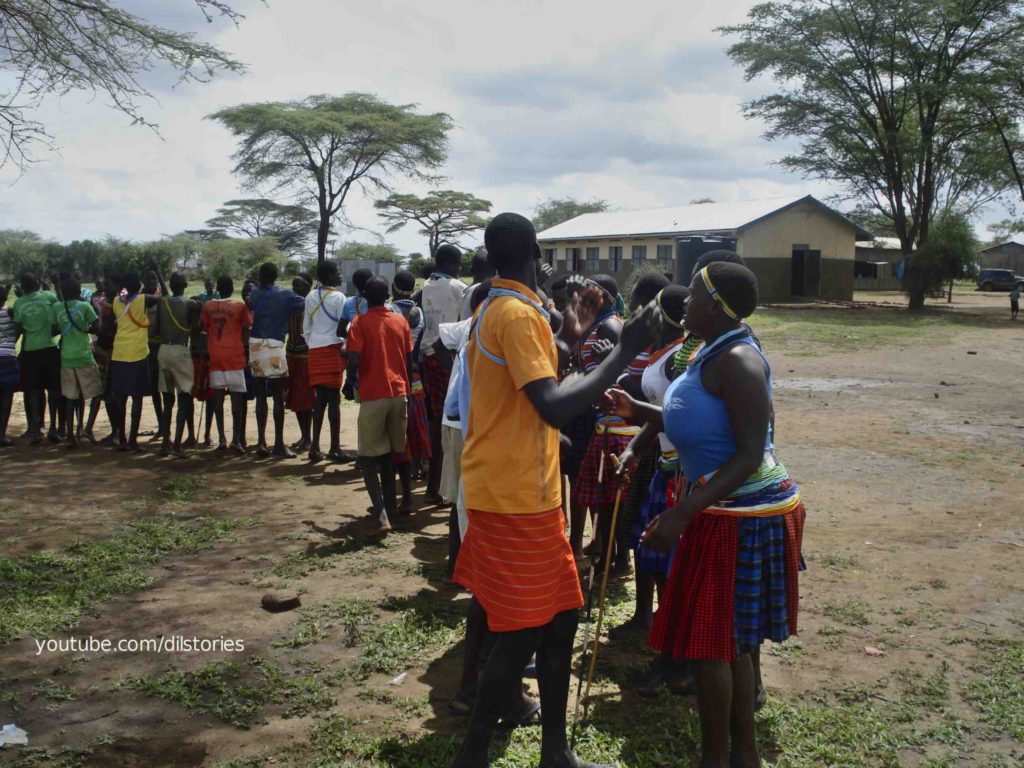 Two rows of dancing facing each other, arranged in a semi-circle, one of boys another of girls, all dressed in traditional karamojong wear