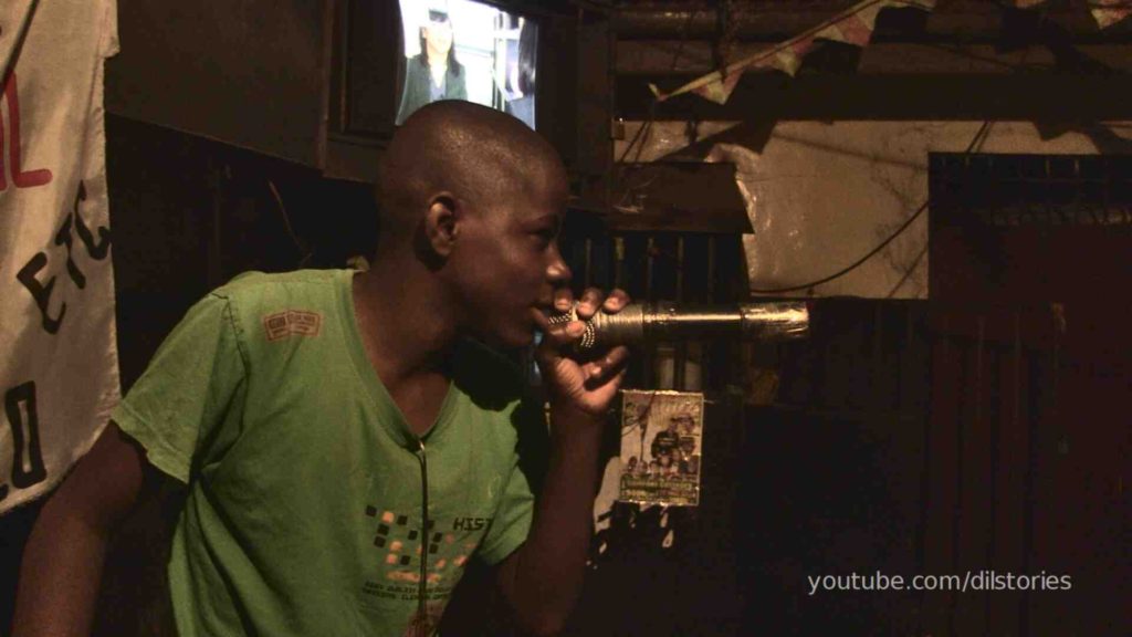 A teenage boy, about nineteen, holding a microphone close to his mouth, rapping on stage in a bar, low lights. TV in the background.