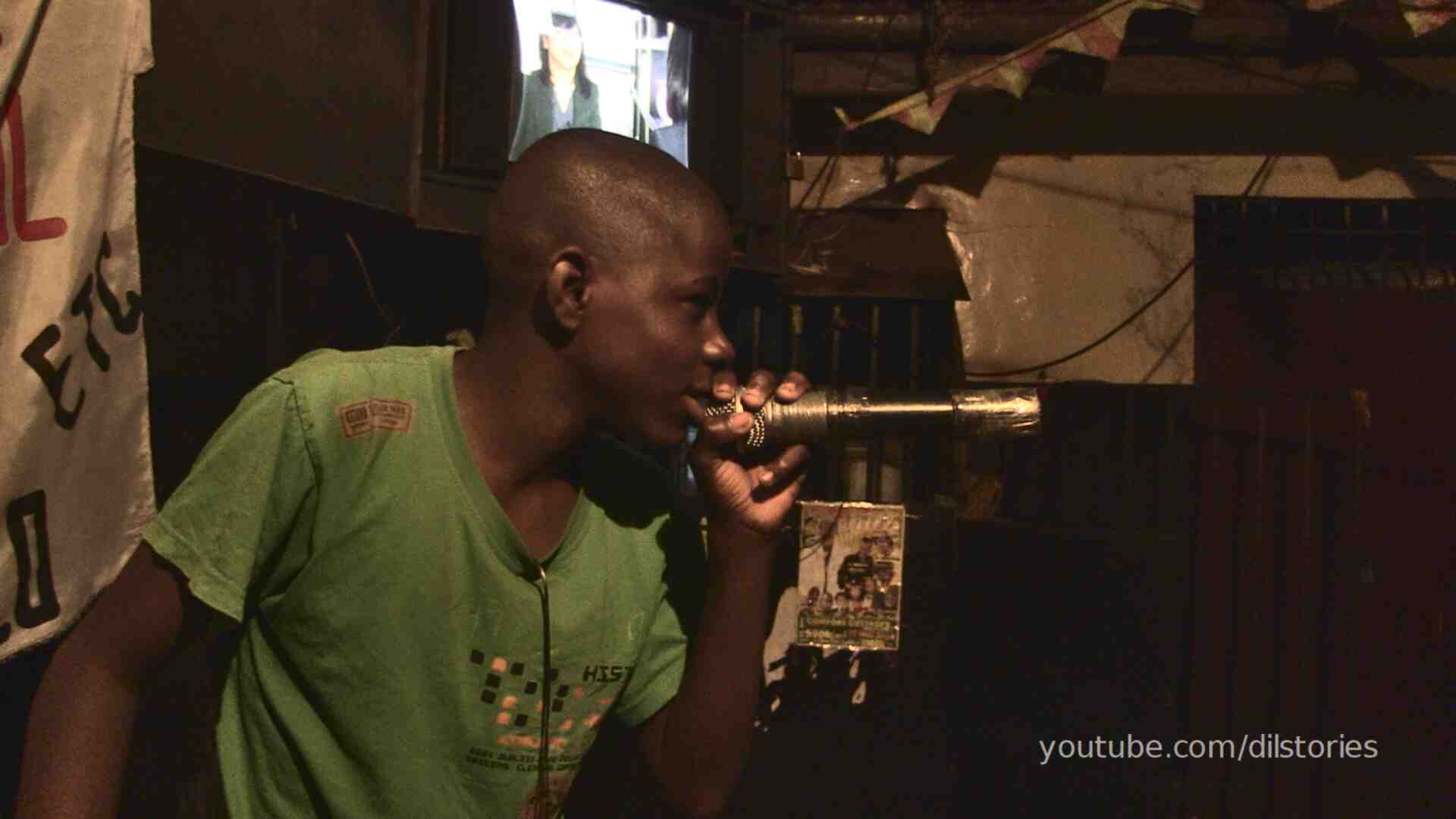 A teenage boy, about nineteen, holding a microphone close to his mouth, rapping on stage in a bar, low lights. TV in the background.