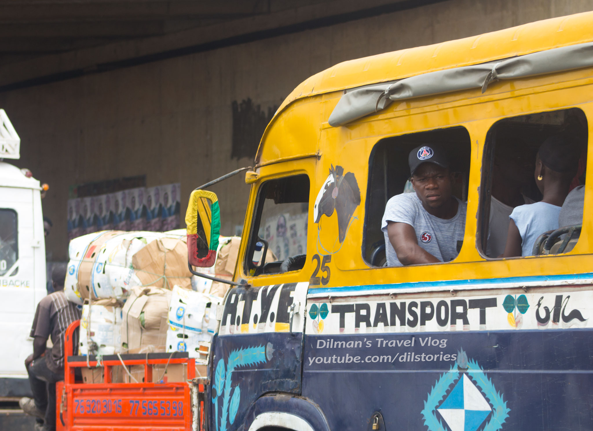 A yellow bus with blue stripes, no glass in the windows, a man staring out of the window.