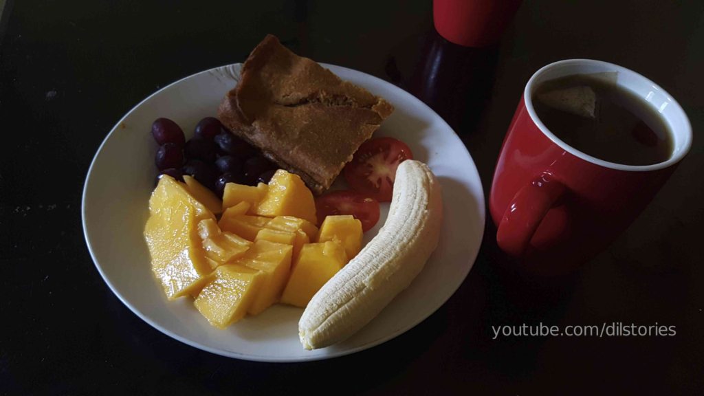 Breakfast on a white plate. Homemade cake. Banana. Pawpaw. Tomatoes. Grapes. Cakes. Red mugs. Tea.