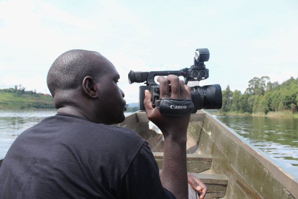 A filmmaker holding a camera, filming, while in a canoe, on a lake.