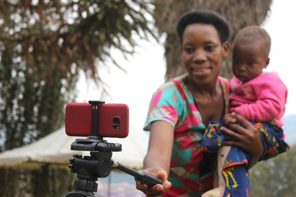 A woman holding a baby, filming with a red smart phone, Samsung S7 in the foreground