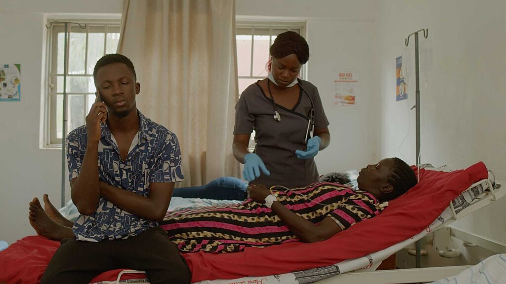 A film scene in a hospital room. A actor sits on a bed, while a woman is sick and lies on the bed, and a nurse towers above the woman.