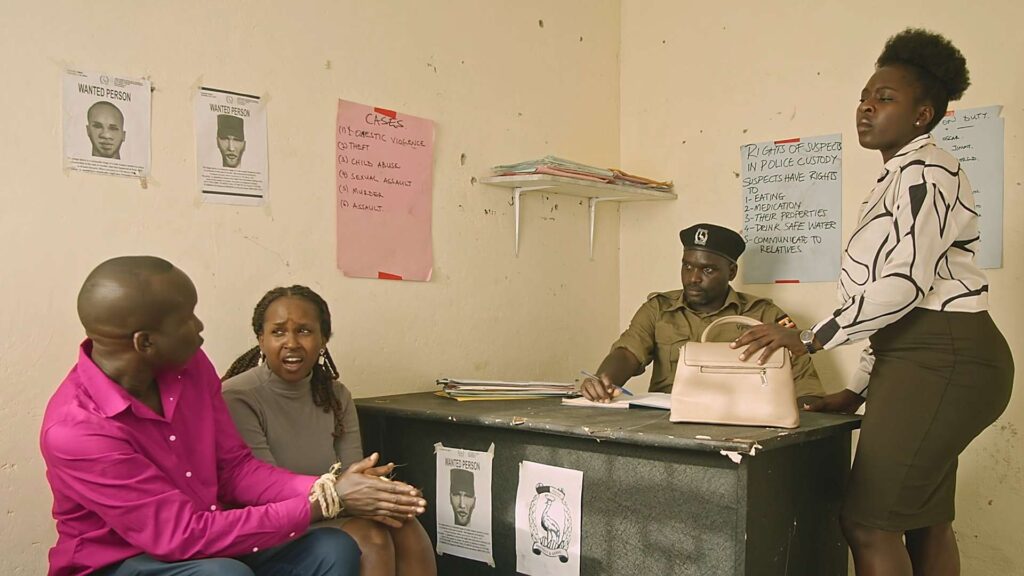 A screenshot of a film scene, in a police station, with a police staring at two suspects, whose hands are bound with ropes, while a woman stands towering above them.