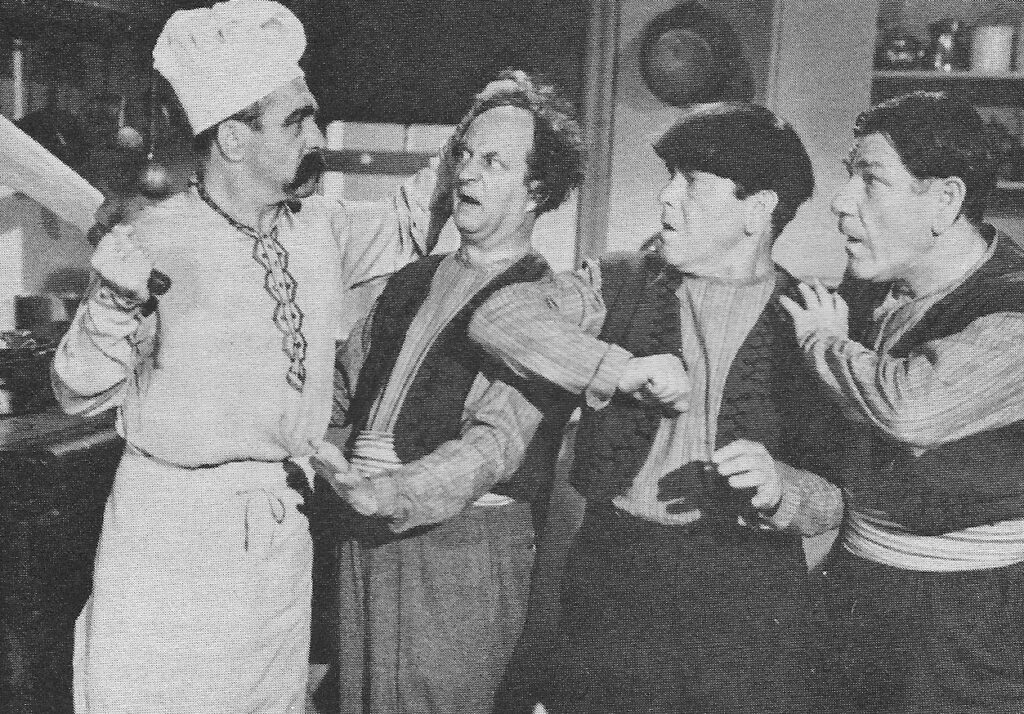 Screenshot of an episode of The Three Stooges, from Malice in the Palace. Three men make clownish faces as they stare at chef, who holds a butcher's knife and spiked moustache.