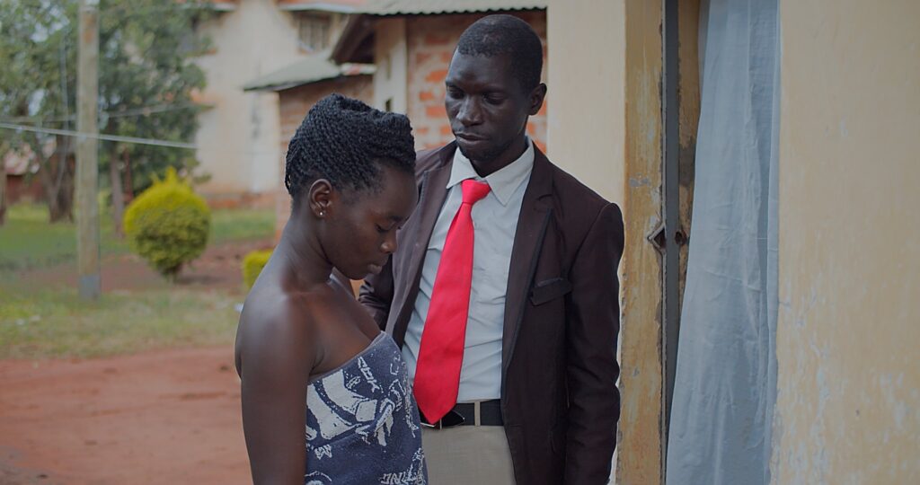 Scene from a film, an African man, middle aged, in a coat and red tie, stares at a younger woman, who is barely dressed with only a towel wrapped around her.