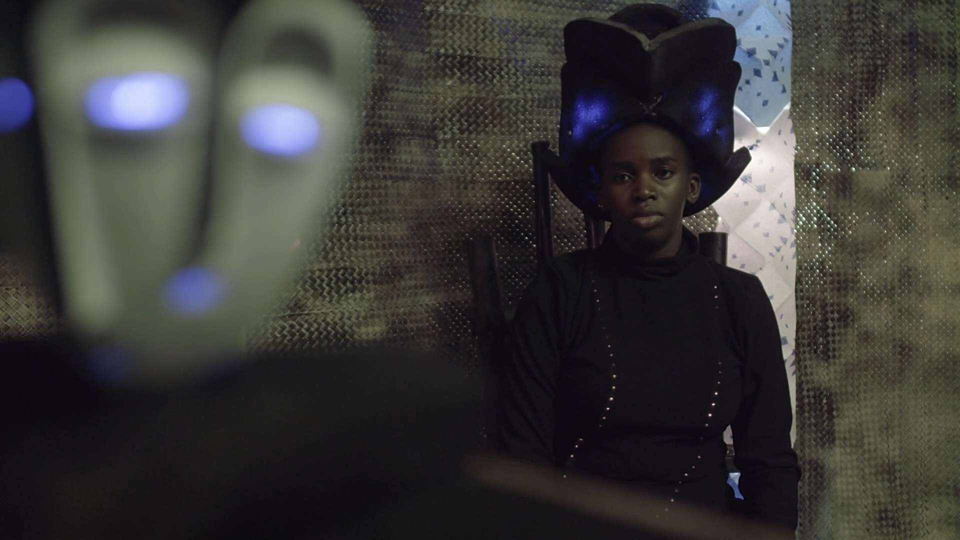A film scene, underlit, a woman in a futuristic hat with blue lights stares at a robot in the foreground, out of focus.
