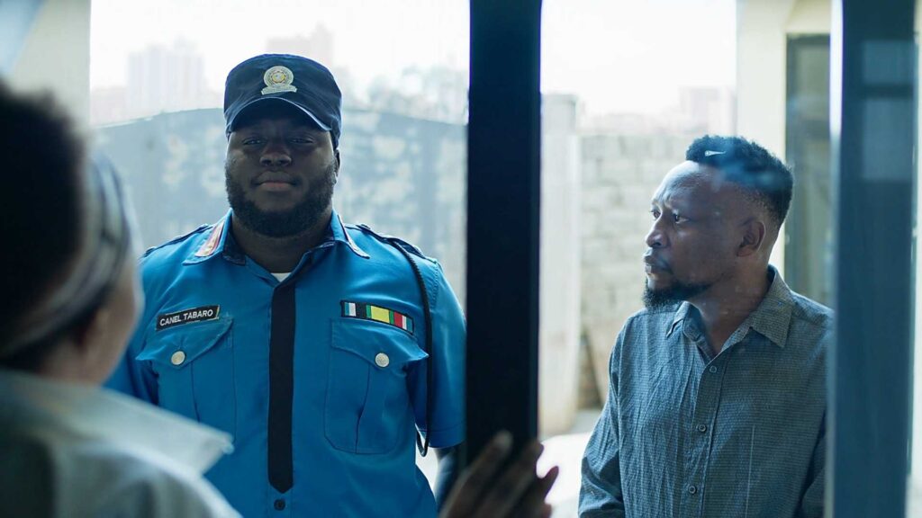 An actor in a police uniform stands at a doorway, while another, more scared individual, stands next to them. They face a woman who is inside the house.