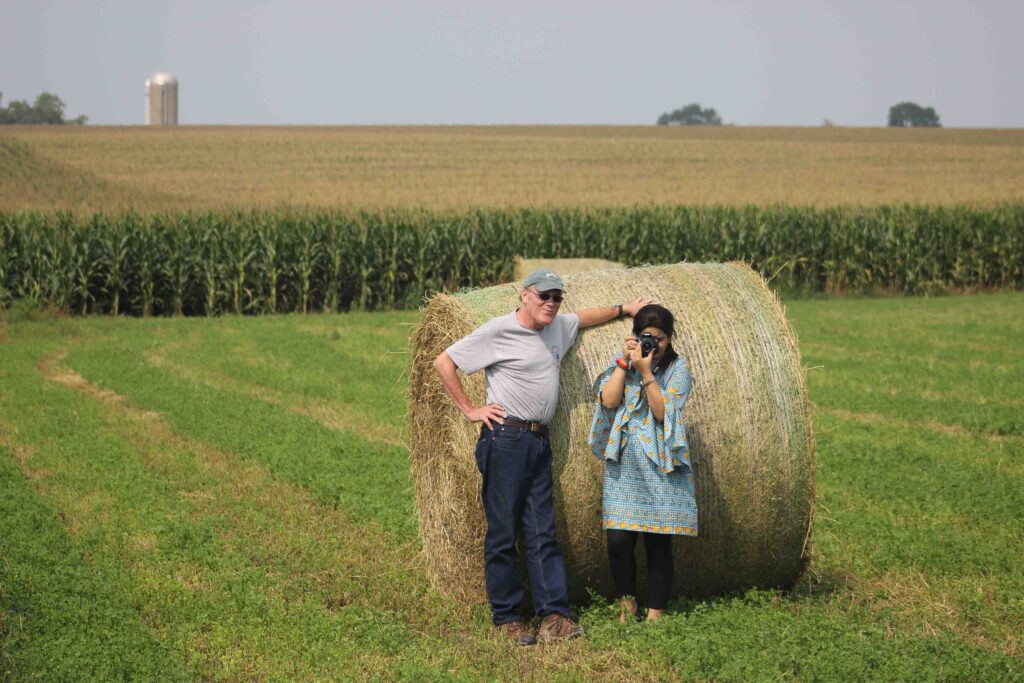 two people pose for a photo at a bale of corn, rolled up, a corn field in the distance