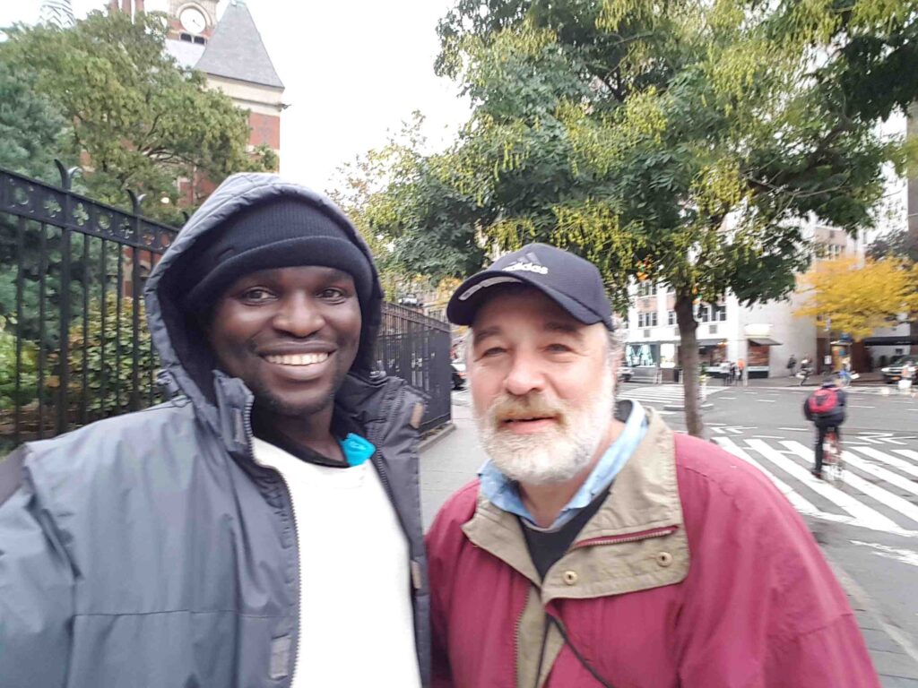 Two men take a selfie in the streets of New York, an african man in a jacket with a hoddie and a white old man with all white beard wearing a cap.