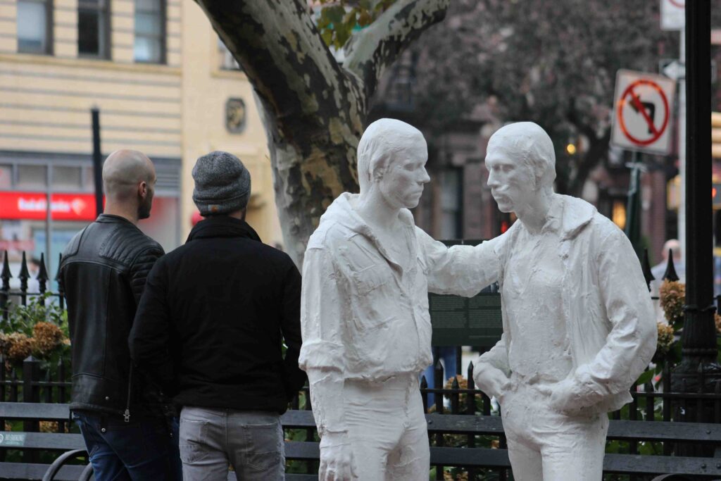 A statue in Stonewall park, of two men, painted white, facing each other, while two people, male-looking, with their backs to the camera, are behind.
