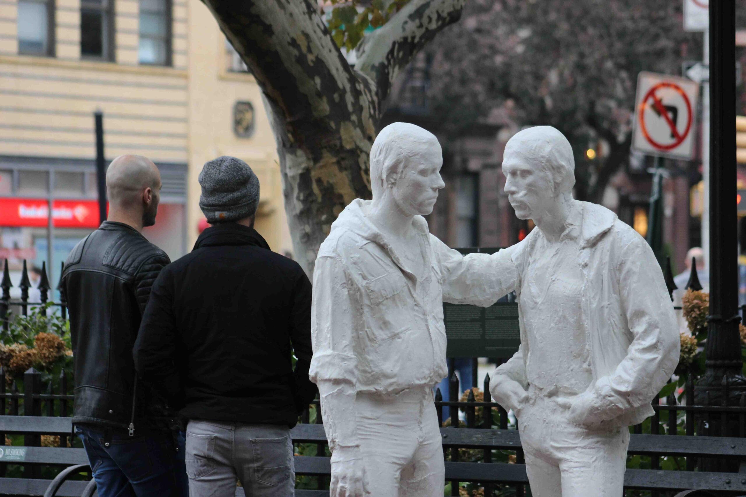 A statue in Stonewall park, of two men, painted white, facing each other, while two people, male-looking, with their backs to the camera, are behind.