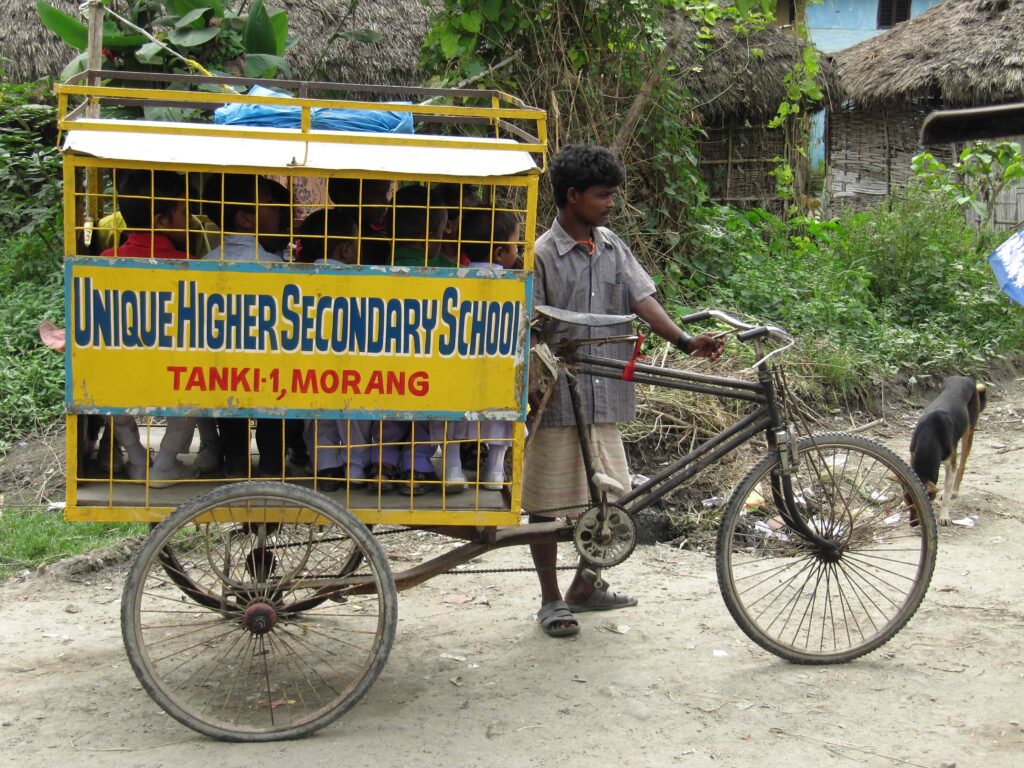 A rickshaw that has been turned into a school bus, ferrying kindergarten children, who are full inside the carrier. It is painted yellow, and has the words Unique Higher Secondary School